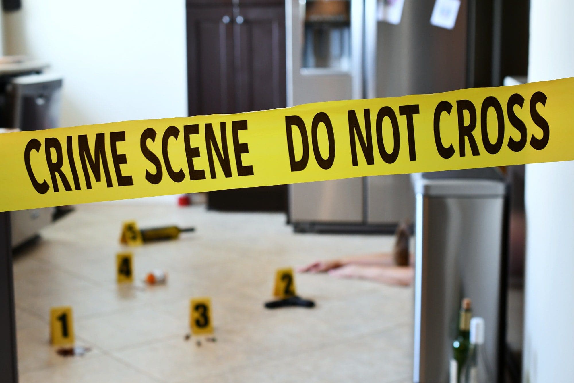 Crime Scene Do Not Cross yellow tape with indoor evidence markers out of focus in the background.