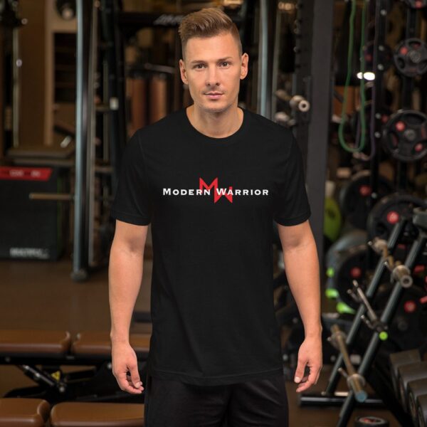 A man wearing a black MW Combative Basic T-shirt stands in front of a gym.