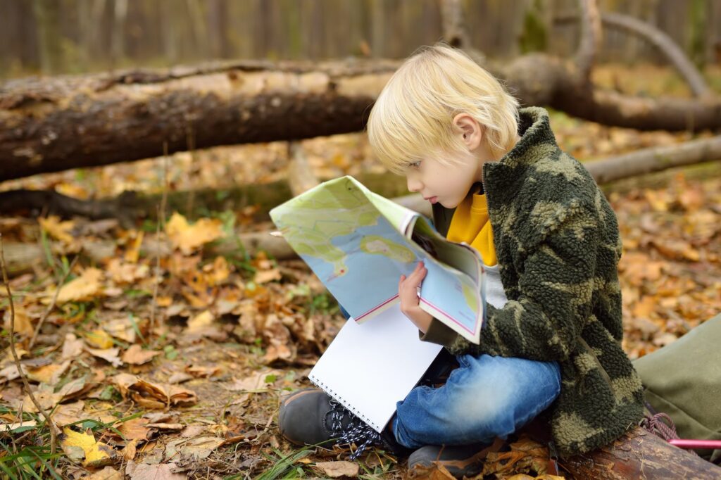 Little boy scout is orienteering in forest. Child is sitting on fallen tree and looking on map