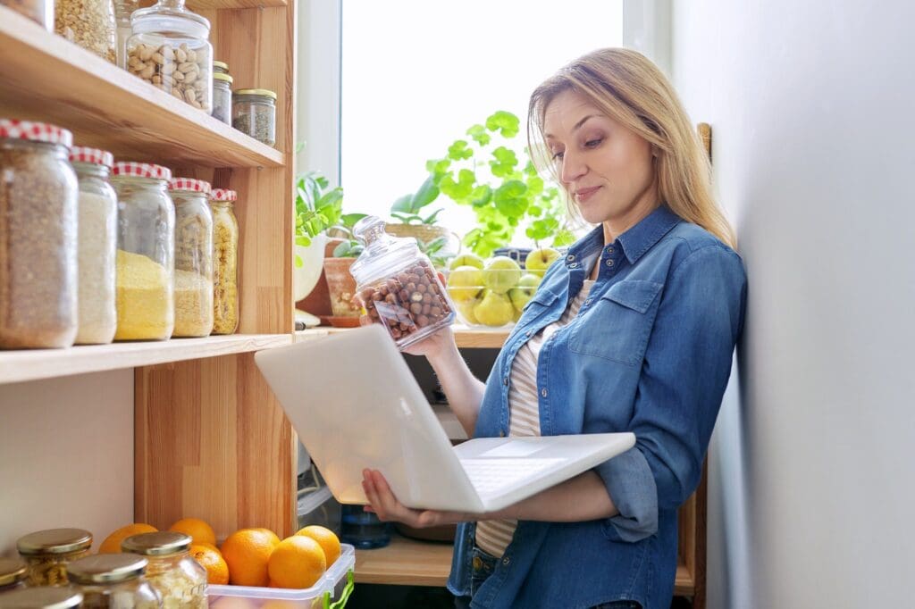 Middle-aged woman in kitchen in pantry with laptop showing cans of food