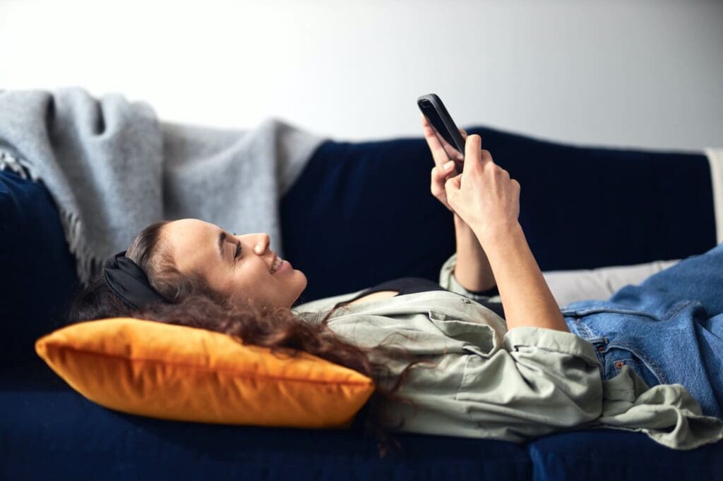 Young Smiling Woman Relaxing At Home Lying On Sofa Checking Social Media On Mobile Phone