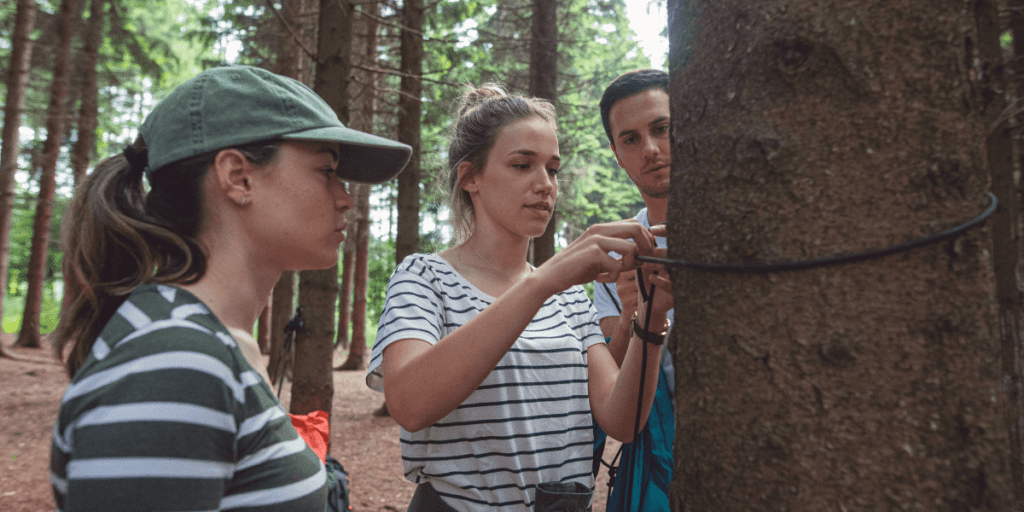 Three young adults, facing financial constraints, are examining a rope tied around a tree in a forested area, possibly engaging in a survival preparedness activity.